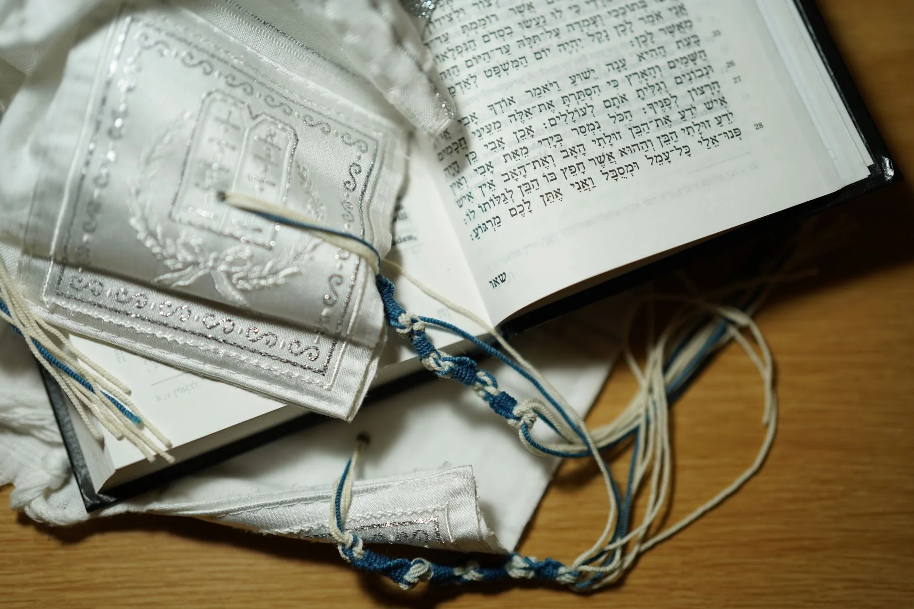 An open Hebrew Bible with a tallit and siddur on a wooden table.