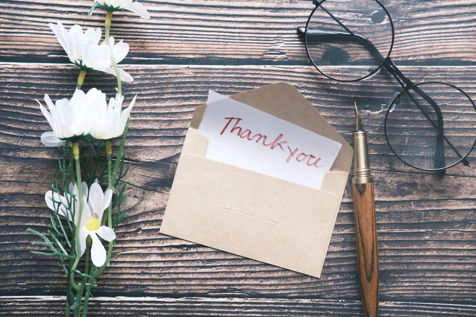 A flat lay of a thank you note in an envelope with daisies and a pen.